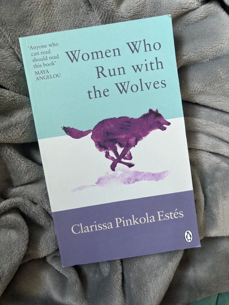 A paperback copy of the book 'Women Who Run with the Wolves' by Clarissa Pinkola Estés, featuring a light blue, white, and purple cover with an illustration of a running wolf, resting on a gray textured blanket.